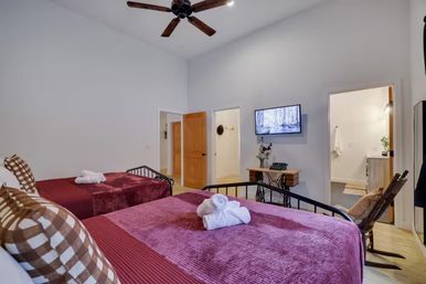 Bright guest bedroom with two metal-frame beds dressed in deep burgundy throws and checkered pillows, rolled towels on the beds, wall-mounted TV, ceiling fan, and open doorway to an en-suite bathroom — cozy vacation rental vibe.