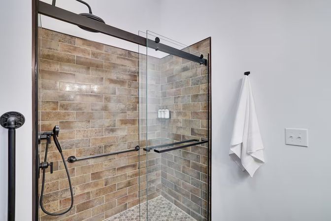 Modern walk-in shower with sliding glass door, brown subway tile walls and hexagon floor tiles, matte-black rain and handheld shower fixtures, glass shelf with dispensers and a white towel hanging on the wall.
