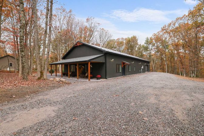 Cozy dark metal-sided barn-style home with a covered wooden porch on a wide gravel driveway, surrounded by trees in orange autumn foliage under a blue sky.