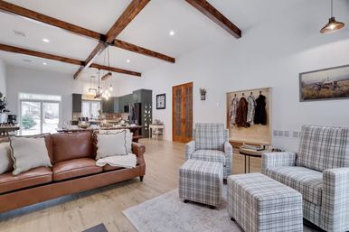 Bright open-concept living room and kitchen with vaulted ceiling and exposed wooden beams, caramel leather sofa, plaid armchairs and ottomans, farmhouse-style coat bench, pendant lights, hardwood floors and sliding glass door to a snowy yard.