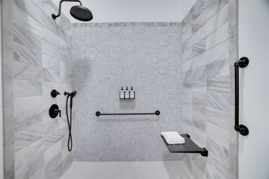 Modern accessible walk-in shower with light gray hex and marble-effect wall tiles, matte black rainfall showerhead and handheld, grab bars and fold-down bench with folded white towels.