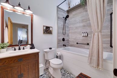 Cozy farmhouse-style residential bathroom with a wooden vanity and mirror, white sink and toilet, tub-shower combo with gray wood-look tile surround, matte black fixtures, pebble mosaic floor and neutral tasseled shower curtain.