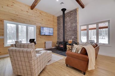 Cozy cabin-style living room with wood-paneled walls, large windows revealing a snowy woodland view, stone accent wall with a black wood-burning stove and stacked firewood, plaid armchairs, leather sofa with knit throw, hardwood floors and wall-mounted TV.