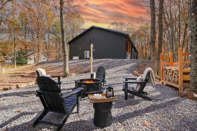 Cozy autumn cabin retreat at sunset with a black modern cabin, gravel patio, Adirondack chairs around a glowing fire pit, stacked firewood and colorful fall trees.
