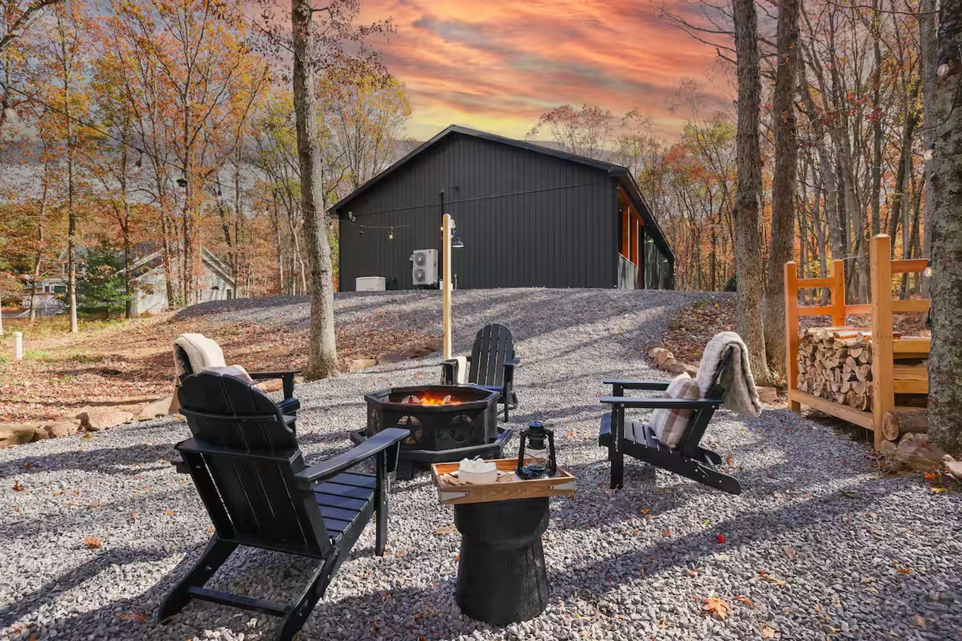 Cozy outdoor fire pit with black Adirondack chairs and blankets on a gravel patio surrounded by autumn woods and a modern cabin at sunset