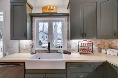 Cozy farmhouse kitchen with a white apron-front sink and black faucet, sage green shaker cabinets, butcher-block countertops, stone tile backsplash, a countertop spice rack, and a window overlooking a snowy yard and bare trees.