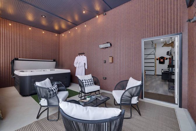 Cozy indoor hot tub lounge with a black spa, four cushioned wicker chairs around a coffee table on a woven rug, string lights overhead, white bathrobes on hooks, and a doorway opening to an adjacent garage/workshop.