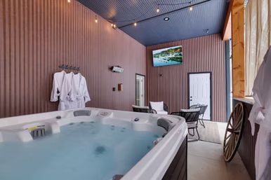 Cozy indoor spa room with bubbling hot tub, white robes on hooks, wicker seating and table, wall-mounted TV, string lights and rustic wooden accents.