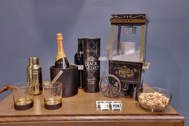 Cozy home bar tabletop with vintage popcorn cart, champagne in an ice bucket, wine bottle and tall liquor tin, gold cocktail shaker, two whiskey glasses, a bowl of mixed nuts and matchboxes.