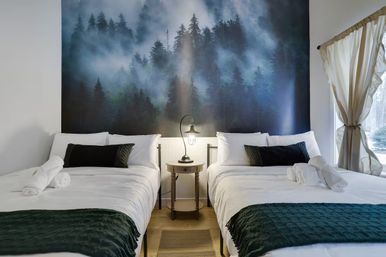 Cozy bedroom with two twin beds in white linens and dark green throws, a central wooden nightstand with a lamp and rolled towels, and a large misty forest mural behind the beds with natural light from a curtained window.