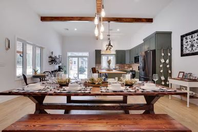 Cozy farmhouse dining room with a long wooden trestle table and benches set for a meal, exposed wood beams and hanging Edison bulbs, open kitchen with sage-green cabinets and stainless appliances, and glass doors revealing a snowy yard.