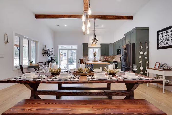 Open-concept modern farmhouse dining room and kitchen with a long wooden farmhouse table set for eight on bench seating, green shaker cabinets, Edison bulb pendant lights, and large windows.