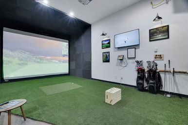 Indoor golf simulator room with large projection screen displaying a virtual course, green artificial turf and hitting mat, golf bags, wall-mounted TVs and framed photos — ready for a virtual round.