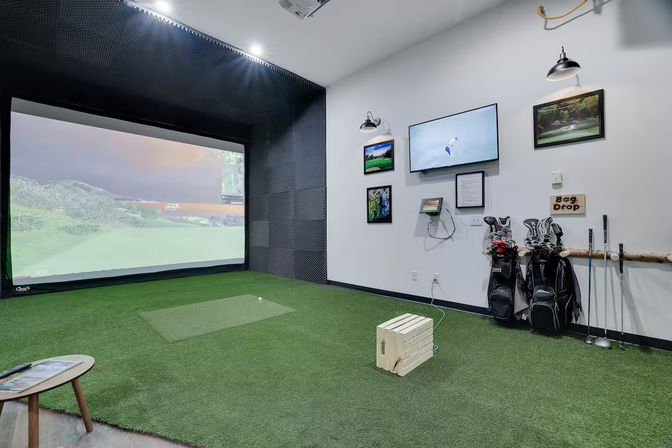 Indoor golf simulator bay with large projection screen showing a virtual course, artificial turf and hitting mat, wooden tee crate, two golf bags with clubs, wall-mounted TV and framed golf photos — ready for a swing.