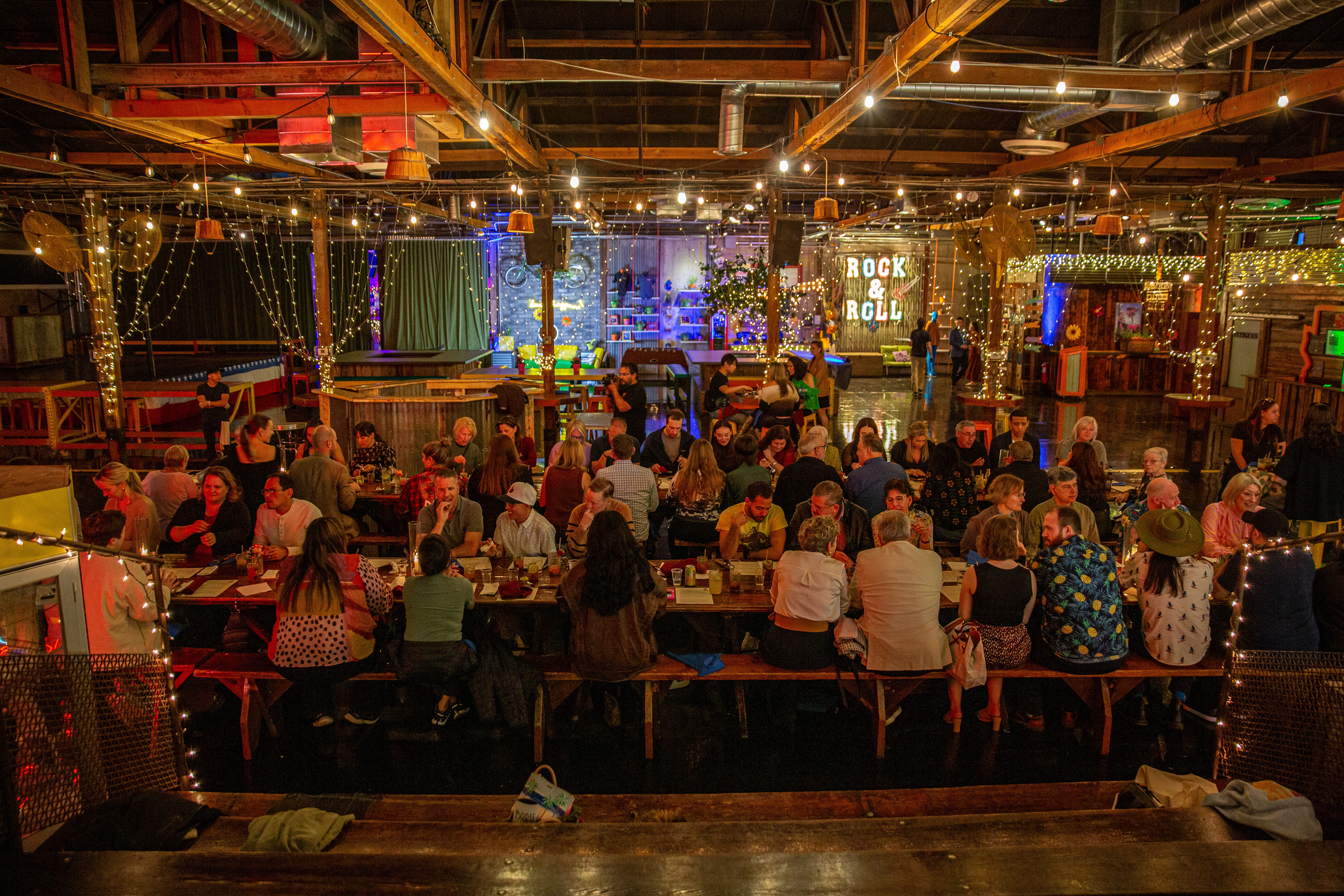 Bustling indoor warehouse-style beer hall with long communal tables, twinkling string lights, exposed wooden beams and a glowing "Rock & Roll" sign.