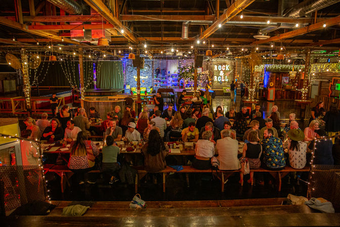 Bustling indoor warehouse-style beer hall with long communal tables, twinkling string lights, exposed wooden beams and a glowing "Rock & Roll" sign.