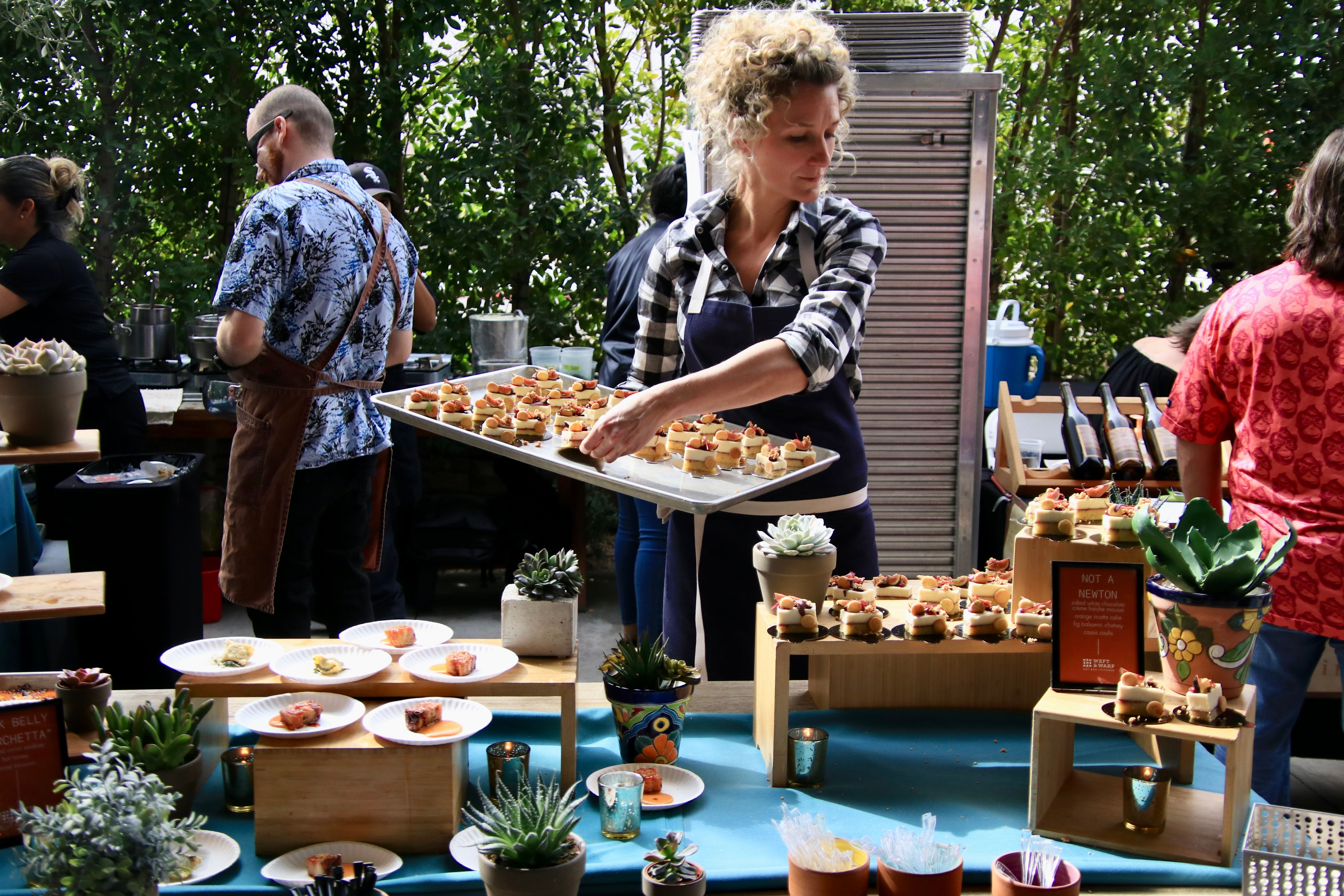 Server arranging bite-sized appetizers on a wooden-riser buffet table with succulents at an outdoor garden-style catering event.