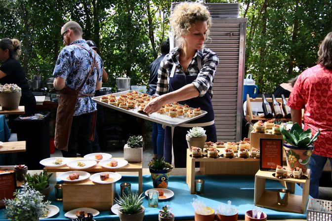 Server arranging bite-sized appetizers on a wooden-riser buffet table with succulents at an outdoor garden-style catering event.
