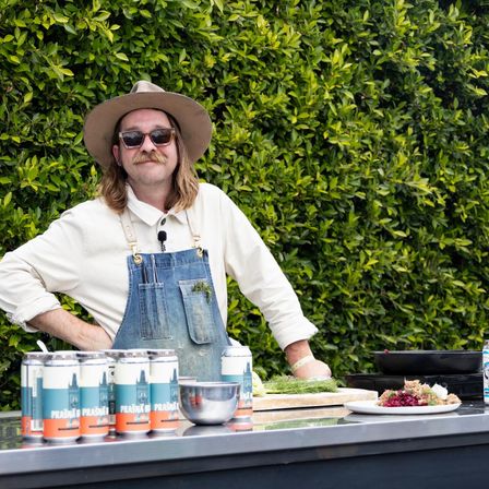 Outdoor chef wearing a wide‑brim hat and denim apron stands at a prep table with craft beer cans, fresh herbs, a mixing bowl and plated dish against a lush green hedge backdrop.