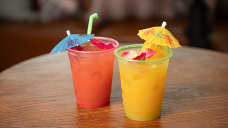 Two festive tropical drinks in plastic cups—one pink and one orange—topped with paper umbrellas, rose petals, ice and straws, sitting on a wooden table.