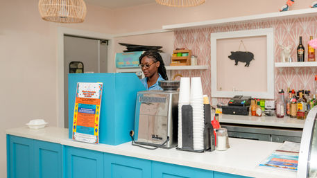 Barista at a bright blue coffee shop counter with an espresso machine, stacked paper cups, condiments and colorful menu, pink patterned wall and pig wall art in the cozy café interior.