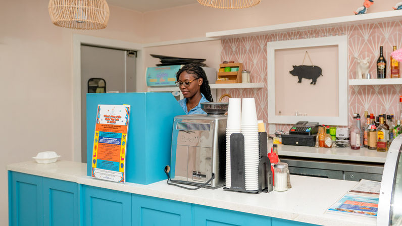 Barista at a bright blue coffee shop counter with an espresso machine, stacked paper cups, condiments and colorful menu, pink patterned wall and pig wall art in the cozy café interior.