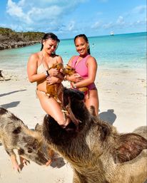 Two women in swimsuits on a sunny tropical white-sand beach holding piglets as curious adult pigs crowd in by clear turquoise water with a boat on the horizon.