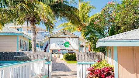 Sunny tropical resort entrance with palm trees, pastel beach cottages, white picket fences, and vibrant tropical flowers
