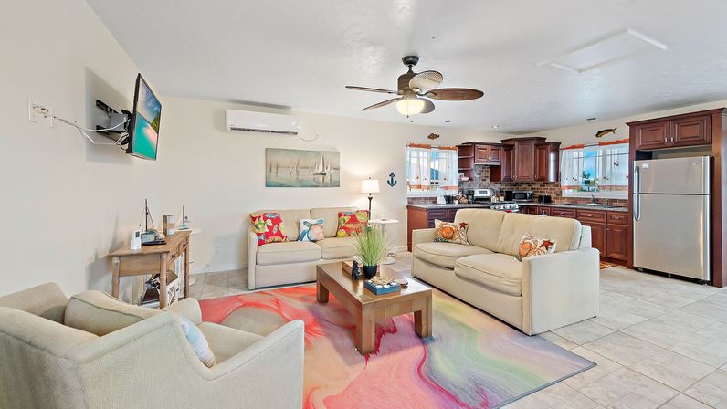 Open-plan coastal-style living room and kitchen with beige sofas, colorful swirled area rug, wooden coffee table, ceiling fan, stainless-steel fridge and dark wood cabinets, beach-themed decor and bright natural light.