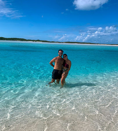 Smiling couple standing in crystal-clear turquoise water at a tropical white-sand beach, sunlit shallow waves and low green islets under a bright blue sky