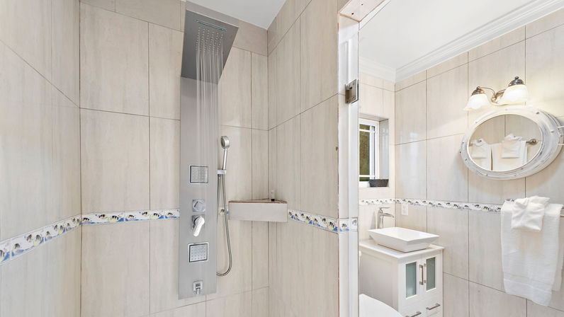 Modern tiled bathroom with stainless steel rainfall shower panel and handheld spray, decorative tile border, white vanity with vessel sink, round mirror and folded towels.