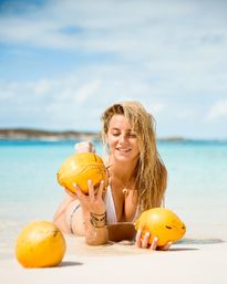 Smiling woman in a white bikini lying on a white-sand tropical beach, holding bright yellow coconuts with turquoise water and blue sky — beachy vibes.