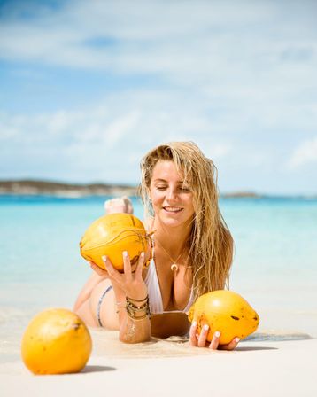 Smiling woman in a white bikini lying on a white-sand tropical beach, holding bright yellow coconuts with turquoise water and blue sky — beachy vibes.