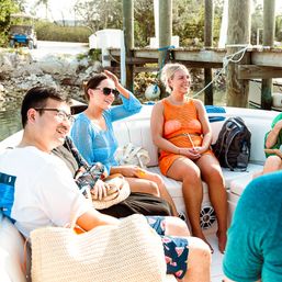 Smiling friends relaxing on a white motorboat tied to a wooden dock, wearing bright beach cover-ups and sunglasses on a sunny summer day with calm water and shoreline trees.
