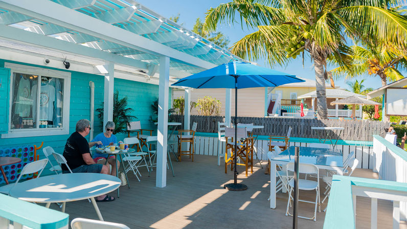 Sunny beachside cafe deck with turquoise siding, blue umbrella and outdoor seating under a palm tree, two people dining on a sunlit wooden patio