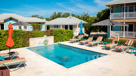Bright tropical poolside scene with a small rectangular swimming pool, striped lounge chairs and colorful umbrellas, pastel coastal cottages and lush greenery under a clear blue sky — vacation rental vibe.