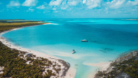 Aerial drone view of a tropical turquoise lagoon and white sandbar with two anchored boats and people wading along a lush, palm‑fringed coastline.