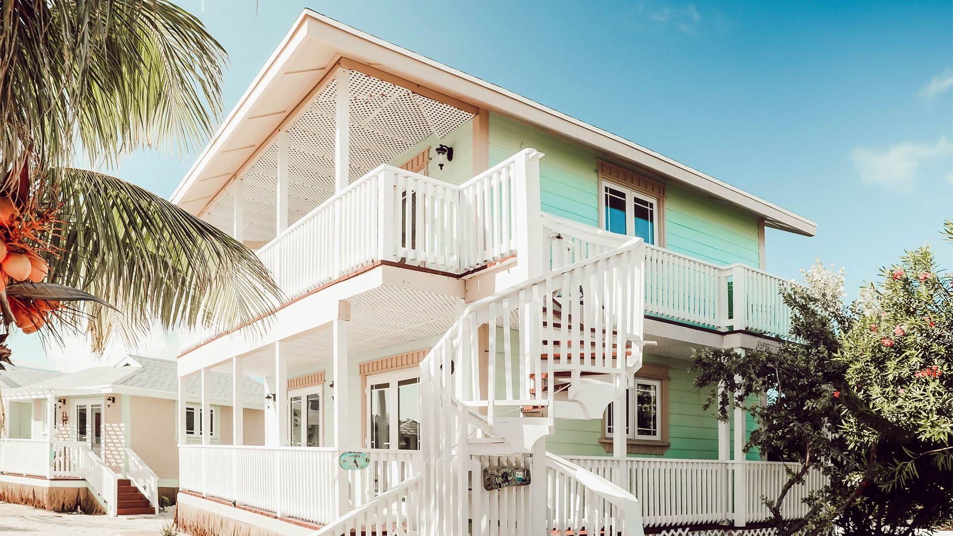 Two-story mint-green tropical beach house with white wraparound porch and exterior spiral staircase, coconut palm in foreground and bright blue sky overhead