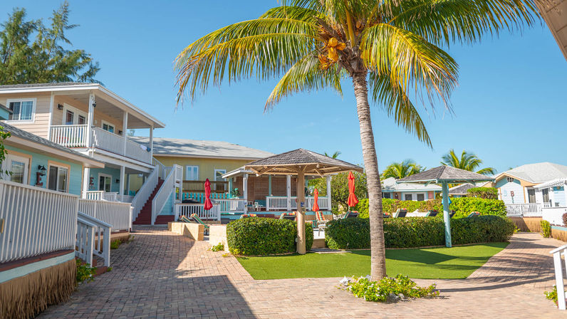 Sunny tropical courtyard at a coastal vacation cottage complex with a tall palm tree, gazebo, lounge chairs and red umbrellas under a bright blue sky