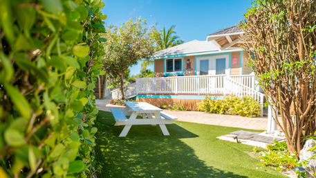 Sunny tropical beach cottage with turquoise trim, white railing and a white picnic table on a manicured lawn framed by hedges and palm trees