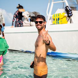 Smiling man in sunglasses and a cap flashing a peace sign while wading in clear turquoise water next to a white motorboat with yellow life jackets on a sunny boat excursion