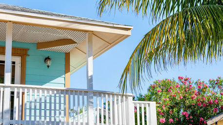 Sunny tropical coastal porch with white railing and turquoise siding, palm frond overhead and pink flowering shrubs against a clear blue sky.