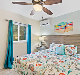 Bright beach-inspired bedroom with starfish-patterned bedding, teal curtains, wooden headboard, bedside table and lamp, ceiling fan, and wall-mounted air conditioner