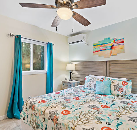Bright beach-inspired bedroom with starfish-patterned bedding, teal curtains, wooden headboard, bedside table and lamp, ceiling fan, and wall-mounted air conditioner