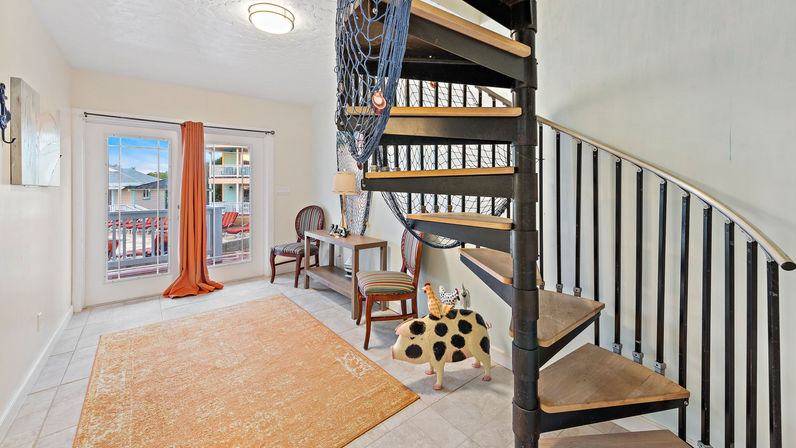 Cozy coastal-style room with black metal spiral staircase, orange area rug on tile floor, sliding glass doors to a balcony with outdoor chairs, nautical netting, striped seating and a playful spotted pig sculpture.