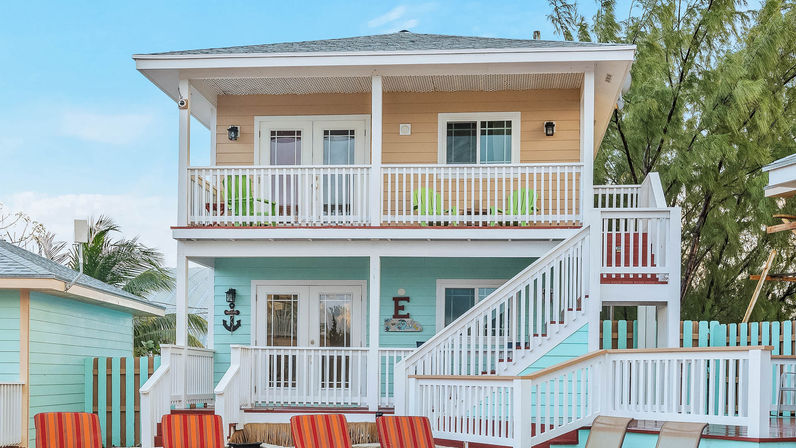 Cheerful two-story pastel coastal cottage with white railings and exterior staircase, turquoise ground floor and sandy-peach upper balcony, bright Adirondack chairs, palm trees and clear blue sky