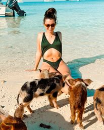 Smiling woman in a green cut‑out swimsuit sitting at a tropical island beach with clear turquoise water while curious piglets explore the sandy shoreline