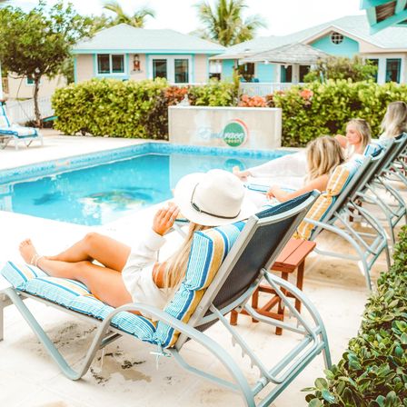 Women lounging on striped pool chairs beside a turquoise pool at a tropical coastal resort, white sun hat, pastel cottages and palm trees in the background.