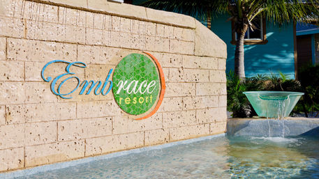 Beige stone wall with a green circular tile logo next to a flowing glass-bowl fountain spilling into a shallow pool, palm tree and turquoise cottage in the tropical resort entrance.