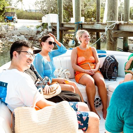 Smiling friends enjoying a sunny boat ride at a wooden dock in a coastal marina, wearing bright summer outfits and sunglasses with beach bags and backpacks onboard.